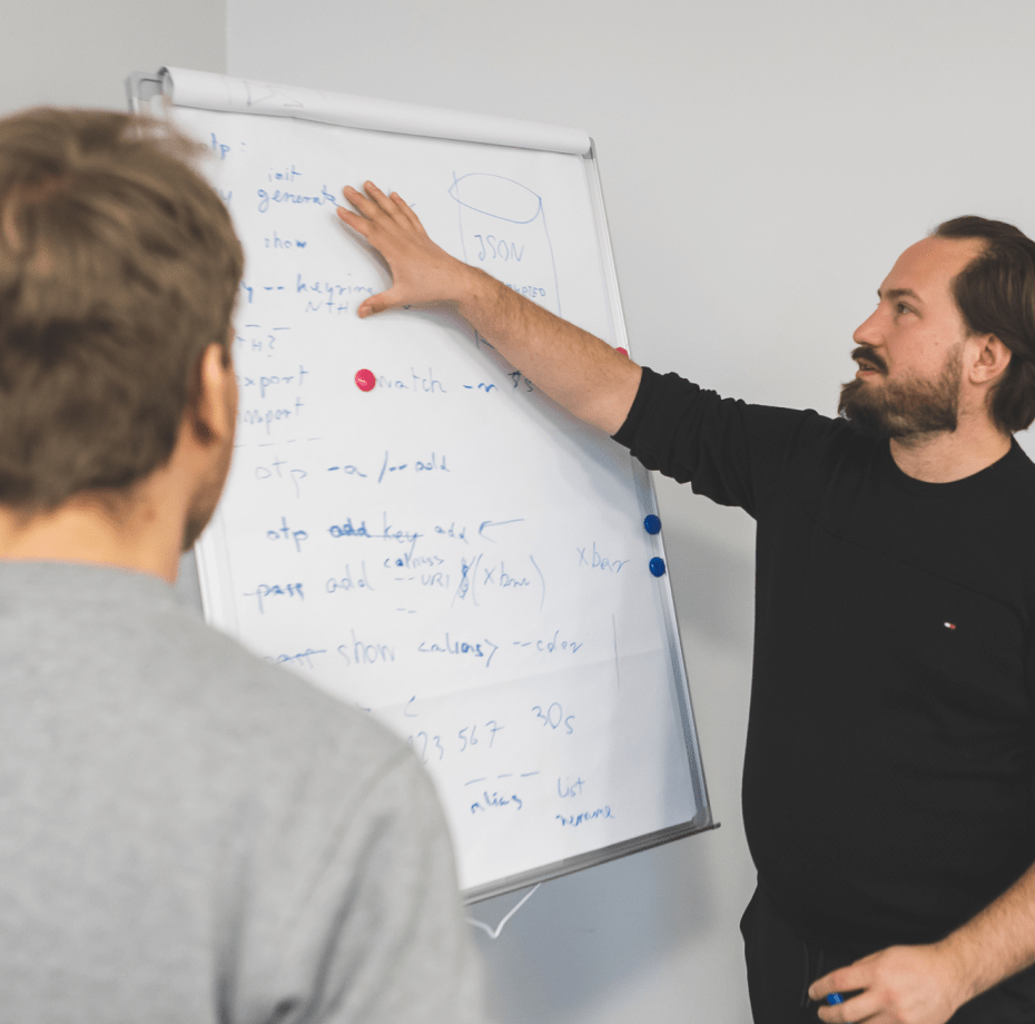 Two programmers standing by a whiteboard, one is pointing to something written on the board