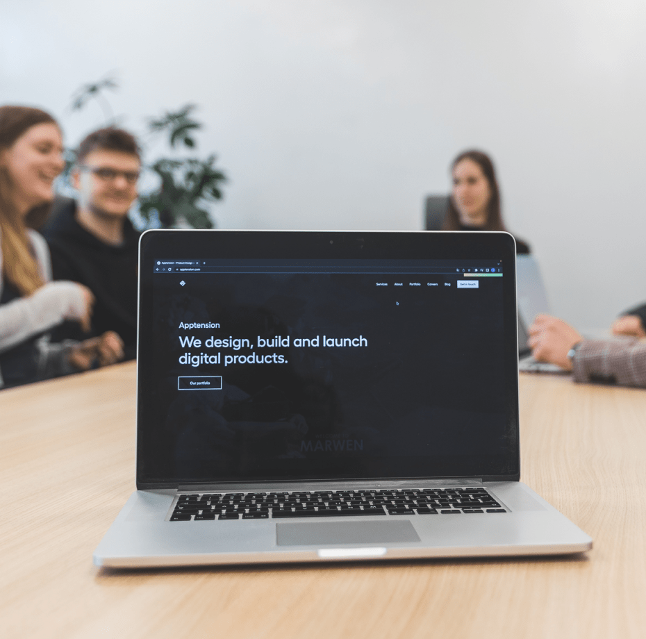A laptop standing on a conference table, in the background there are three people working at Apptension